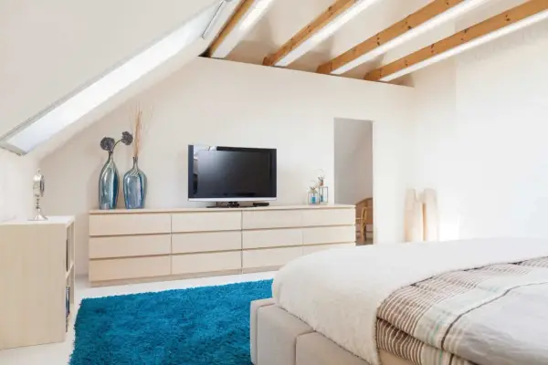 Schlafzimmer im Dachgeschoss, ideal zum Dachboden ausbauen. Helle Farben, Holzbalken und ein blauer Teppich schaffen Gemütlichkeit.